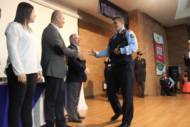 Eine Gruppe von Menschen auf einer B√ľhne, einer in Uniform mit einer M√ľtze, eine Frau in einem wei√üem Hemd, mit einem Banner, Lautsprecher, Vorh√§ngen, Tisch und Bildschirm im Hintergrund.