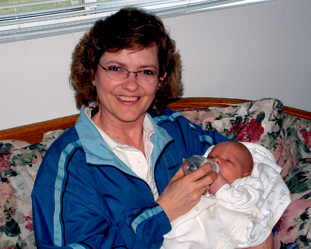 Eine Frau in einer blauen Jacke sitzt auf einem Sofa und füttert ein Baby, mit einem Fensterladen und einer Wand im Hintergrund.