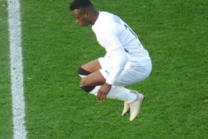 Ein Mann in weißer Uniform und Fußballstiefeln kniet auf einem Fußballfeld, mit einer weißen Linie auf der linken Seite des Bildes.