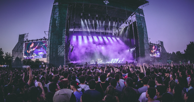 Eine große Menschenmenge auf einem Musikfestival schaut auf einem hell erleuchteten Bühne mit Instrumenten und Bildschirmen, die vor einem Gebäude, Bäumen und einem klaren blauen Himmel aufgebaut sind.