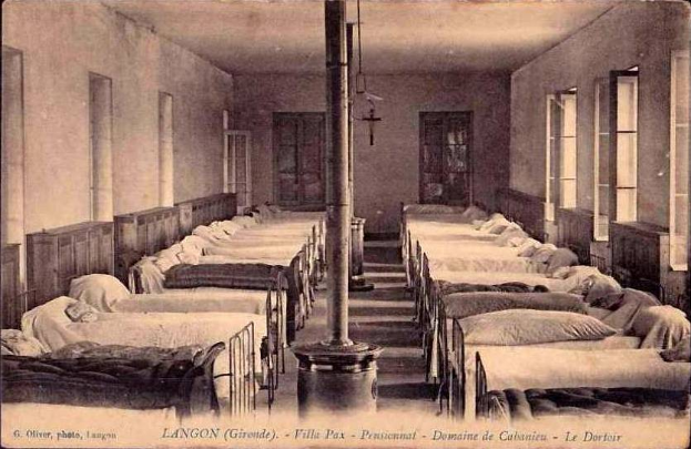 Ein altes Schwarz-Weiß-Foto eines Krankenhauszimmers mit vielen Betten, Kissen, Decken, einem Pfahl, Fenstern, Türen und einem Kreuz an der Wand, mit Text unten lesend "Langon, Gironde - Fille Pax - Pensionnat - Domaine de Galenien - Le Dortour".