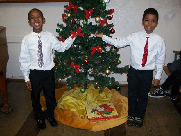 Ein geschm├╝ckter Weihnachtsbaum steht in einem Raum mit einer Kiste und anderen Gegenst├Ąnden auf dem Boden, einem h├Âlzernen Tisch links, einem Stuhl rechts und zwei Jungen in weißen Hemden, die l├Ącheln und den Baum halten.