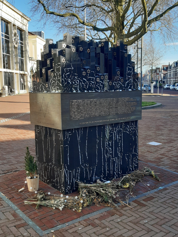 Denkmal in einem Stadtplatz für Holocaust-Opfer, mit einem Baum davor, einer Topfpflanze und trockenen Blättern auf dem Boden, umgeben von Gebäuden, Fahrzeugen, Pfählen, Bäumen und Himmel.
