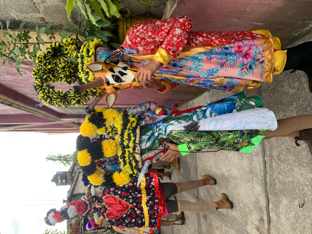 Eine Gruppe von Menschen in bunten Kostümen steht vor einem Gebäude und hält verschiedene Gegenstände, mit Bäumen, Gebäuden und einem klaren blauen Himmel im Hintergrund bei einem Faschingsumzug.