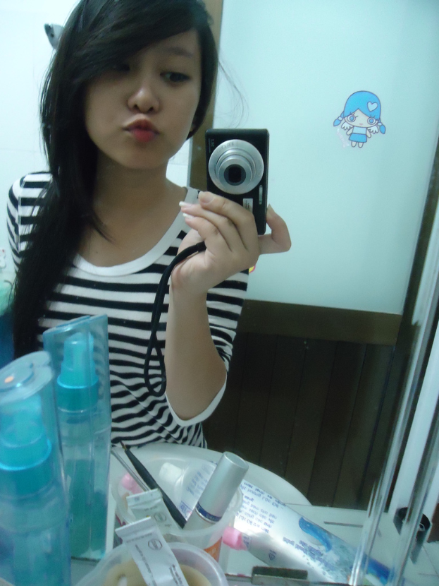 Eine Frau steht vor einem Schminktisch mit Kosmetikartikeln und hält eine Kamera in der Hand, im Hintergrund ist eine Tür zu sehen.