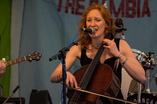 Eine Frau in einem schwarzen Kleid spielt Geige vor einem Mikrofon und einem Ständer, mit einem Banner und einer Band im Hintergrund.