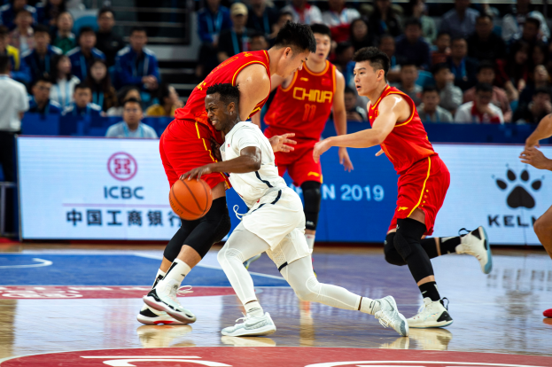 Eine Gruppe von Männern, die Basketball in einer Stadion spielt, wobei einer einen Ball hält und Zuschauer im Hintergrund sitzen.