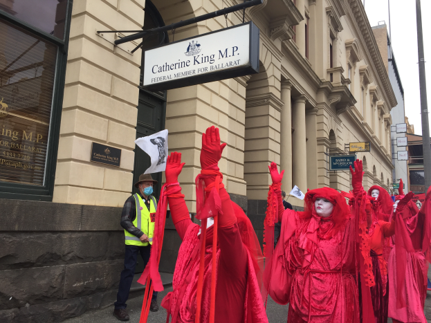 Eine Gruppe von Menschen in roten Kostümen geht eine Straße entlang während eines Ballarat Faschingsumzugs, mit einer Person in einer grünen Jacke und einem Hut, die ein Papier in der linken Hand hält, mehreren Brettern, die an einem Pfahl vor einem Gebäude befestigt sind, und dem Himmel oben im Bild.