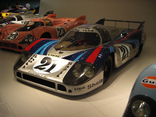 Ein Porsche 917K Le Mans Rennwagen in einem Museum ausgestellt, umgeben von anderen Sportwagen, mit Menschen in der Nähe und einer Wand im Hintergrund; ein Textschild ist links sichtbar.