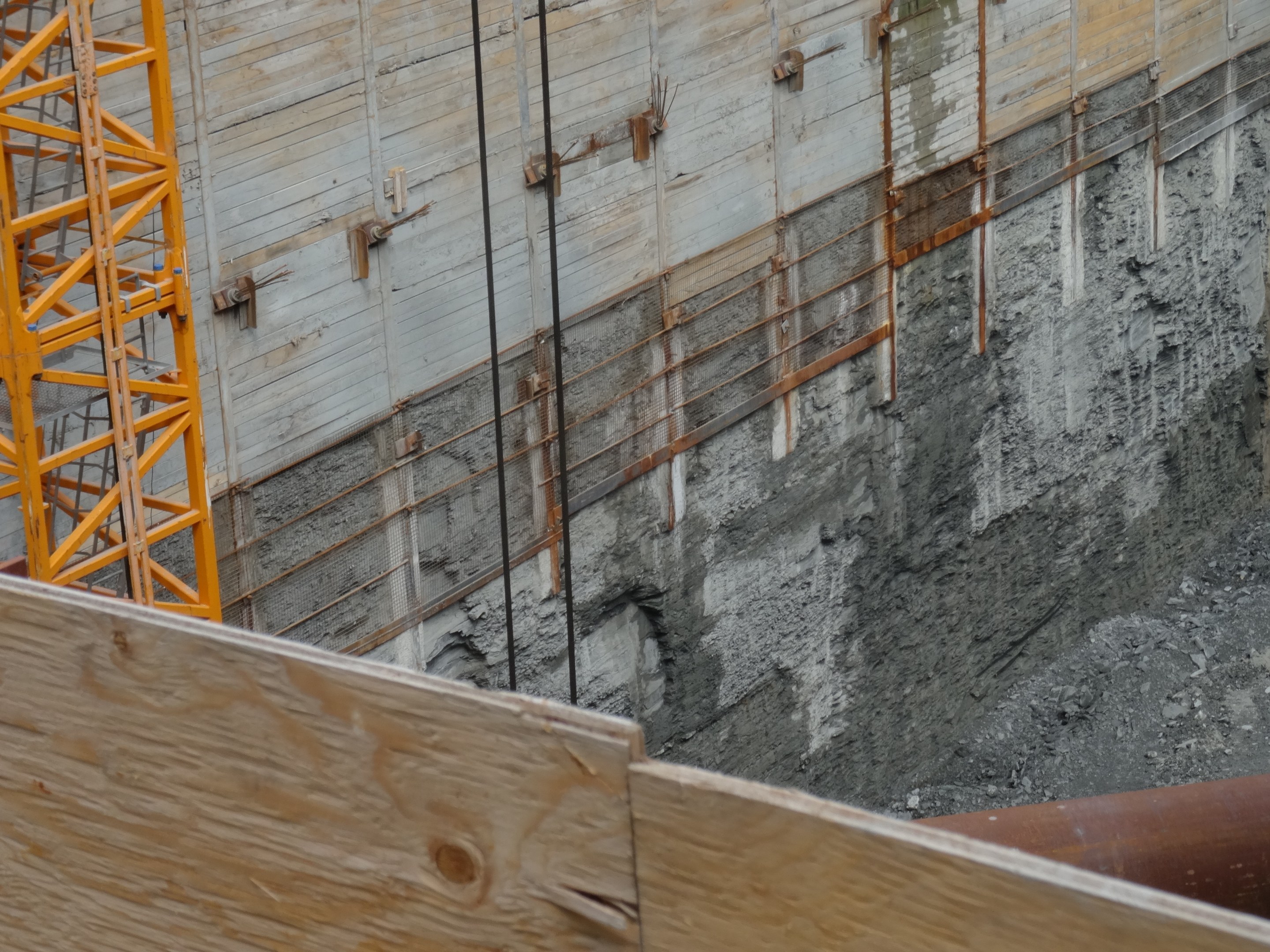 Baustelle mit einem Kran auf der linken Seite, einer betonüberzogenen Wand im Hintergrund, einem hölzernen Objekt und einer Rohrleitung unten.