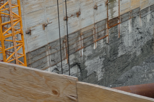 Baustelle mit einem Kran auf der linken Seite, einer betonüberzogenen Wand im Hintergrund, einem hölzernen Objekt und einer Rohrleitung unten.