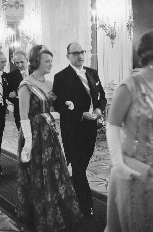 Ein Mann in einem schwarzen Blazer, weißem Hemd und schwarzen Hosen sowie eine Frau in einem eleganten Kleid gehen einen Flur entlang, umgeben von einer Gruppe von Menschen, die auf dem Boden stehen, mit ein paar an der Wand angebrachten Lampen, die eine gemütliche Atmosphäre schaffen.