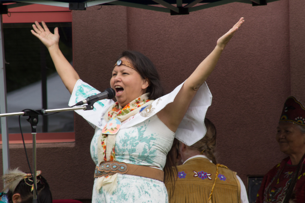 Eine Frau in einem weißen Kleid steht auf einer Bühne, spricht in ein Mikrofon, mit einer Wand und einem Eisenrahmen im Hintergrund.