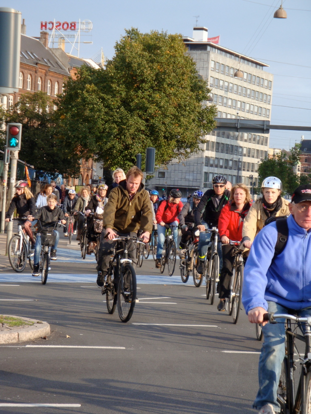 Menschen auf Fahrrädern auf einer Straße mit Pfosten, Bäumen, Gebäuden und dem Himmel im Hintergrund.