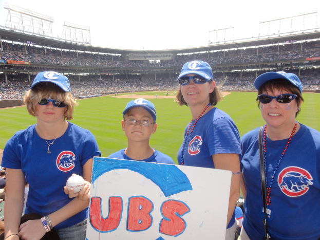 Drei Frauen mit Kappen und Brillen, ein Junge mit Brille und Kappe, der einen Ball und ein Schild hält, in einem Stadion mit Zuschauern und Feld dahinter, unter einem Himmel mit einigen Lichtern.