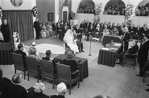 Schwarzes und weißes Foto einer Hochzeitszeremonie in einem großen Raum mit sitzenden und stehenden Gästen, Tischen mit Mikrofonen und Papieren, einer Wand mit einem Fotorahmen, Blumenbouquets, einem Vorhang und einem Podium mit einem Mikrofonständer.