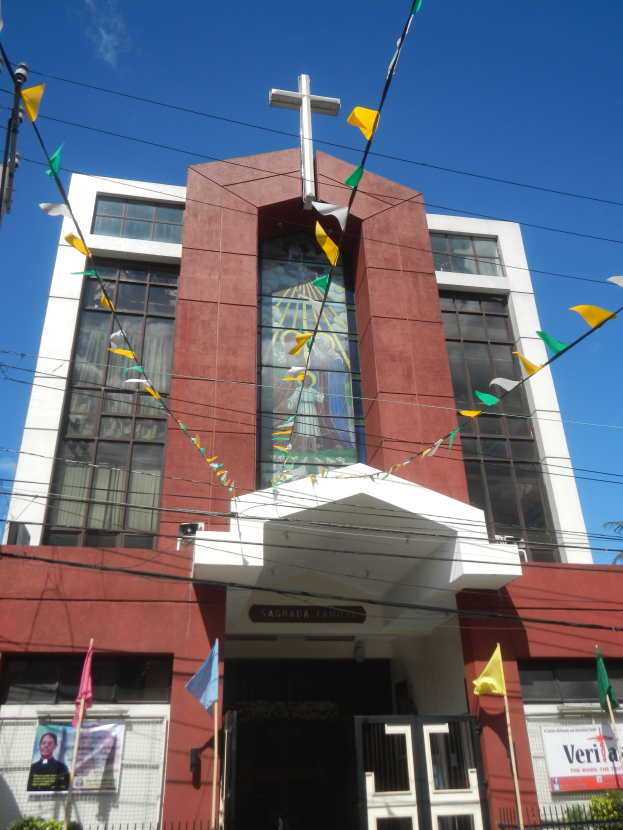 Eine Kirche mit einem Kreuz vorne drauf, umgeben von Fahnen, Pflanzen, einem Metallzaun, einer Fahne mit Text, einem Straßenpfahl, Drähten, einem Baum und einem bewölkten Himmel.