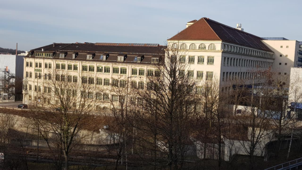 Ein großes, mehrfenstriges Gebäude, der ehemalige Sitz der Deutschen Bundeskanzlei, steht in einer von Bäumen umgebenen Stadt, umgeben von Fahrzeugen, Pfosten und einem Geländer, mit einem bewölkten Himmel im Hintergrund.