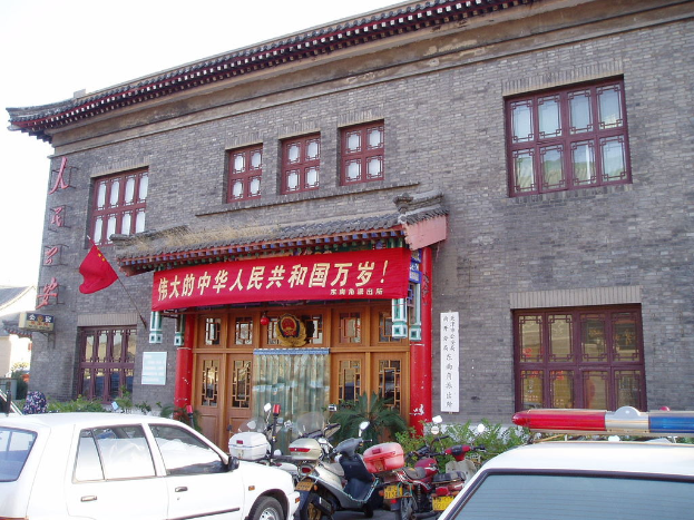 Eine Straßenszene mit Häusern, Pflanzen, Fenstern, Türen, Fahrzeugen, Motorrädern, Trägern, Plakaten, Bannern mit Text, dem Himmel und einer Flagge.