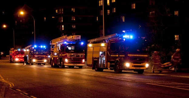 Feuerwehr-Löschfahrzeuge sind nachts auf der Straße unterwegs, mit Gebäuden und Laternen im Hintergrund und zwei Personen, die rechts gehen.