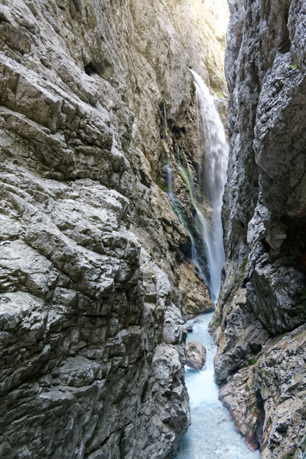 Kleiner Wasserfall, der an zerklüfteten Felsen in einem steinigen Tal hinabstürzt, umgeben von saftig grünen Hügeln mit kristallklarem Wasser bei strahlendem Sonnenschein.