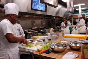 Eine Gruppe von Köchen in einer Küche, die Essen auf einem Schneidbrett zubereiten, wobei ein Koch im Vordergrund in einer weißen Schürze und einer weißen Mütze steht und ein Messer hält, umgeben von verschiedenen Utensilien, Schüsseln, Gemüse, einem Mobiltelefon, Flaschen und anderen Gegenständen, mit einem Fernseher und Deckenleuchten im Hintergrund.