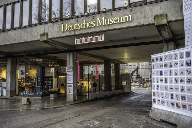 Außenansicht des Deutschen Museums in Berlin, das eine große Glasfassade mit Säulen, ein Namensschild, ein Informationsschild mit Bildern und Text auf der rechten Seite und umliegende Gebäude, Bäume und Himmel zeigt.