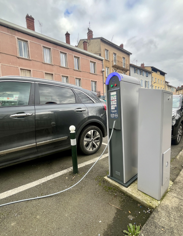 Elektrisches Auto-Laden an einer Straßenstation mit Gebäuden, Laternenmasten und einem bewölkten Himmel im Hintergrund.
