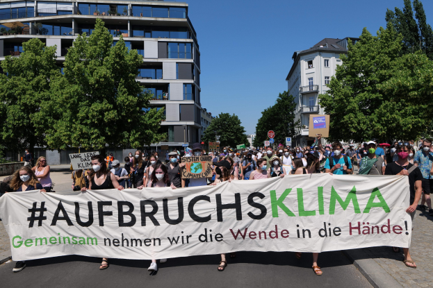 Eine Gruppe von Menschen mit Masken hält ein Schild mit der Aufschrift "Aufbruchsklima" vor einem Gebäude, umgeben von Bäumen und einem klaren Himmel.