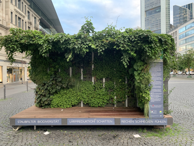 Ein grünes Gebäude mit Efeu an der Seite, umgeben von Bäumen, Fahrzeugen, Pfählen, Schildern und anderen Gebäuden unter einem bewölkten Himmel, Teil der Staufbilter Biodiversität in Berlin, Deutschland.