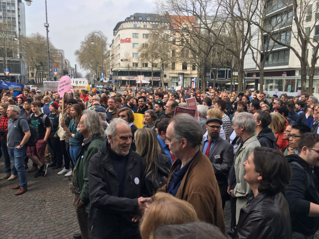 Große Gruppe von Menschen vor einer Menge mit Schildern, Fahrzeugen, Bäumen, Laternenpfählen, Gebäuden und einem bewölkten Himmel während einer Demonstration gegen Regierungssparmaßnahmen in Berlin, Deutschland.