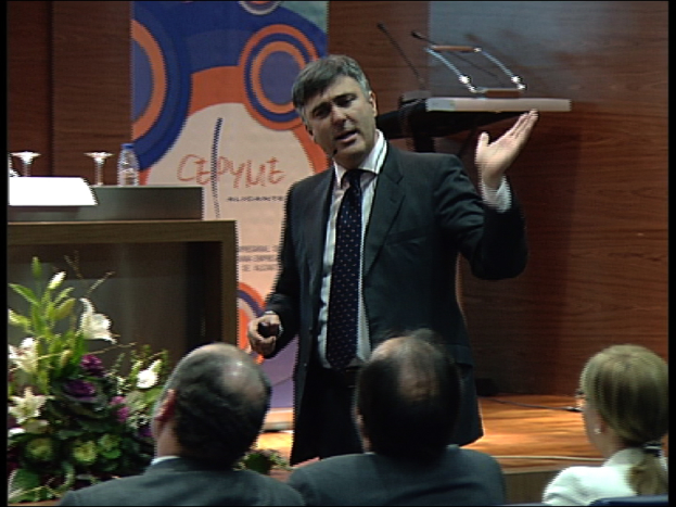 Ein Mann in einem schwarzen Blazer steht an einem Podium mit Mikrofonen, spricht in ein tragbares Mikrofon, mit einer Blumendekoration und einem Tisch mit einem Buch und einer Wasserflasche in der Nähe, während drei Personen auf Stühlen vor ihm sitzen, vor einer braunen Wand und einem bunten Schild mit Text.