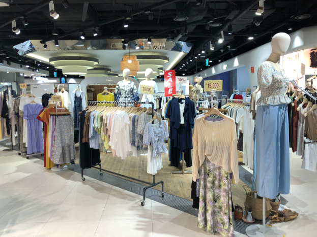 Ein Bekleidungsgeschäft mit Schaufensterpuppen in Kleidern, Kleidung an Bügeln, Texttafeln, Deckenbeleuchtung und verschiedenen Gegenständen auf dem Boden, die darauf hinweisen, dass es ein Modegeschäft ist.