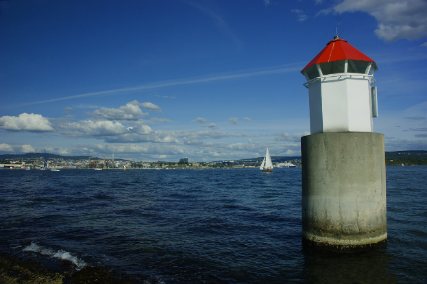 Boote auf dem Meer mit einem Leuchtturm im Vordergrund, Gebäude, Berge und Himmel im Hintergrund.