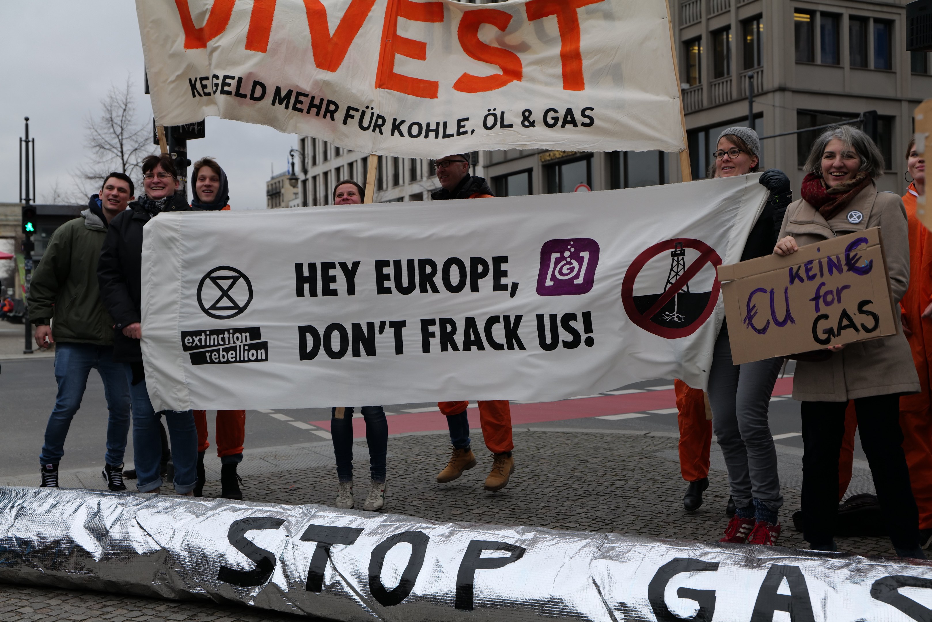 Gruppe von Menschen mit einem "Stoppt Gas"-Schild auf der Straße, die gegen die Europäische Union protestiert. Im Hintergrund sind Gebäude, Bäume, Pfosten, Verkehrszeichen und ein klarer blauer Himmel zu sehen.