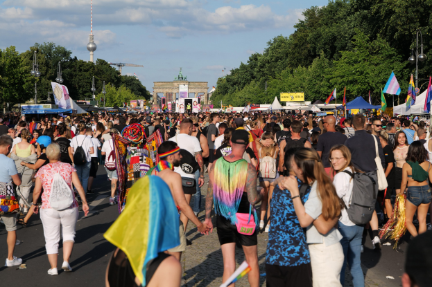 Eine große Menschenmenge marschiert auf einer Straße in Berlin, Deutschland, viele tragen Taschen und halten Fahnen, mit Zelten an den Seiten und Bäumen, Laternenmasten, einem Bogen, einem Turm und Wolken im Hintergrund.