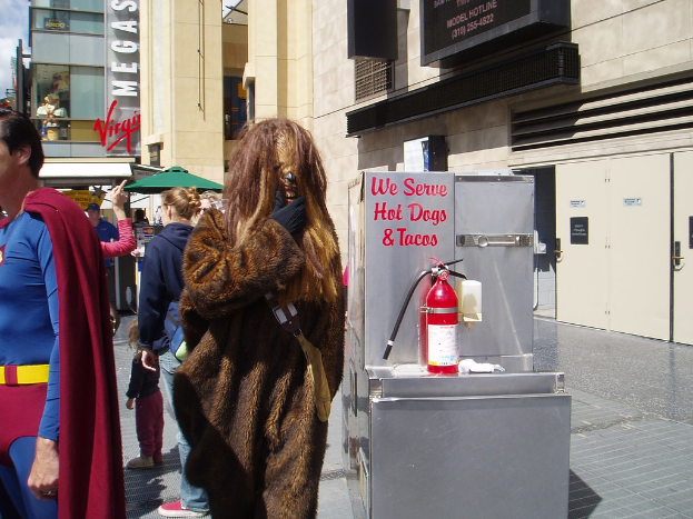 Eine Menschenmenge, einige in Kostümen, versammelte sich vor einem Gebäude mit sichtbarer Beschriftung und Türen, mit einer Box und einem Feuerlöscher in der Nähe.
