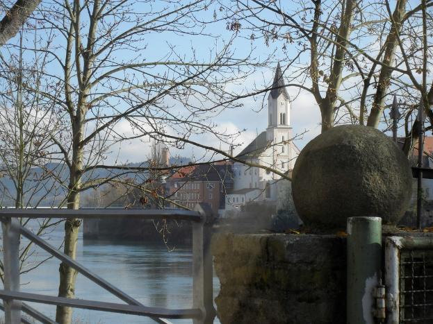 Ein hoher Pfeiler steht vor Gebäuden mit Fenstern, Bäumen mit Zweigen, einem Tor und fließendem Wasser.