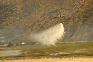 Ein Hubschrauber fliegt über ein bergiges Gebiet und lässt Wasser auf ein Feuer darunter fallen, mit Sand und Wasser unten und Hügeln, Bäumen und Fahrzeugen im Hintergrund.