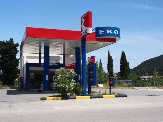 Eine Tankstelle mit einem Schild "Eko" umgeben von Pflanzen mit Blumen, einem Fahrrad, Schildern mit Text, Polen, einer Straße, Bäumen, Häusern, Hügeln und einem bewölkten Himmel im Hintergrund.