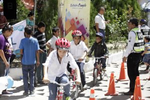 Gruppe von Kindern auf Fahrrädern auf einer Straße mit Verkehrskegeln, einige tragen Helme, andere stehen in der Nähe, mit einer Fahne, Bäumen und Gebäuden im Hintergrund.