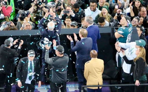 Philadelphia Eagles-Spieler feiern mit dem Vince Lombardi Trophy nach dem Sieg im Super Bowl, einige tragen Mützen, andere halten den Trophy oder werfen Konfetti, mit einem Geländer und Fotografen im Hintergrund.