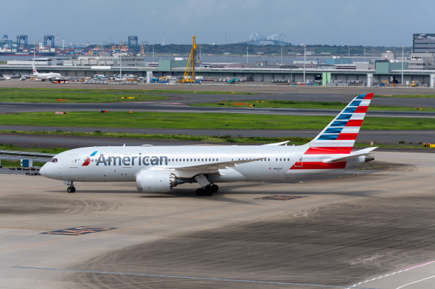 Ein American-Airlines-Flugzeug steht auf der Rollbahn eines Flughafens, umgeben von Gras, Fahrzeugen, Gebäuden, Polen, Kränen und anderen Gegenständen, mit Bäumen und einem klaren blauen Himmel im Hintergrund.