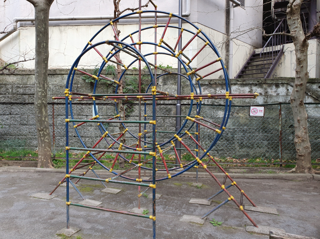 Ein Spielplatz mit einer großen Metallstruktur in der Mitte, umgeben von Bäumen und einer Wand mit Maschendrahtzaun, mit Stufen mit Geländern, die zu einem Gebäude mit Fenstern und Rohren führen, und einer Tafel an einem Pfahl, alles auf einem grasbewachsenen Boden.