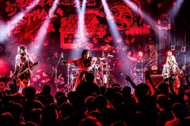 Ein Live-Auftritt der japanischen Rockband Korn auf der Bühne, mit Bandmitgliedern, die Gitarren spielen und in Mikrofone singen, einer beleuchteten Wandmalerei im Hintergrund und einem Publikum unten.