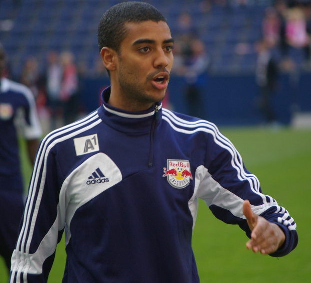 Ein Mann in einer blauen Jacke steht auf einem Fußballfeld, umgeben von einer Gruppe von Menschen, mit einem entschlossenen Gesichtsausdruck.