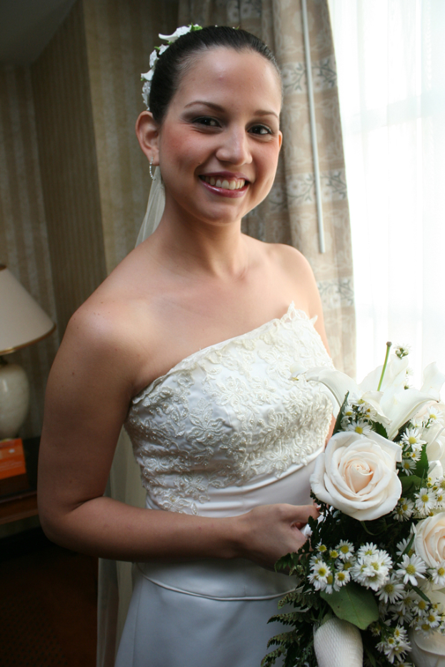 Eine Braut steht lachend und hält einen Strauß in der Hand, mit einem Vorhang und einer Wand im Hintergrund und einer Lampe auf einem Tisch links.