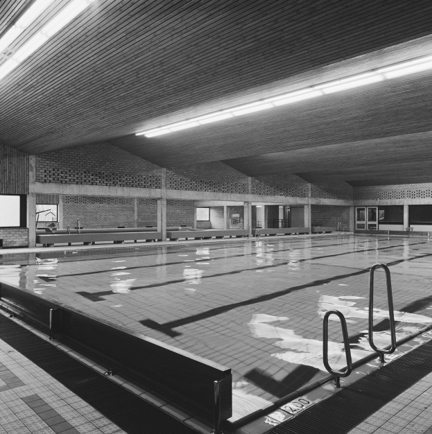 Ein Hallenbad in einem großen Gebäude mit gefliestem Boden, Metallstangen, Wänden, Fenstern und Deckenleuchten, dargestellt in Schwarz-Weiß.