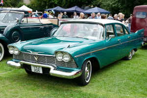 Vintage-Autos auf Gras geparkt mit Menschen dahinter, Zelte im Hintergrund, Bäume oben und eine Flagge oben links.