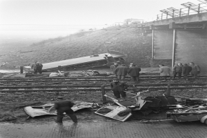 Ein Schwarz-Weiß-Bild einer Zugunfallstelle auf den Schienen mit einer Gruppe von Menschen um das Wrack herum, einem Fahrzeug im Vordergrund und einer Brücke, Pfählen, Drähten und dem Himmel im Hintergrund.
